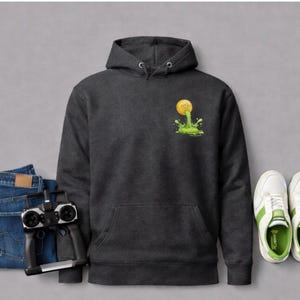 Peut inclure: Un sweat &agrave; capuche gris fonc&eacute; avec une poche avant et un graphisme d'une balle jaune avec de la slime verte. L'image comprend &eacute;galement un jean bleu, une t&eacute;l&eacute;commande et des baskets blanches avec des accents verts.