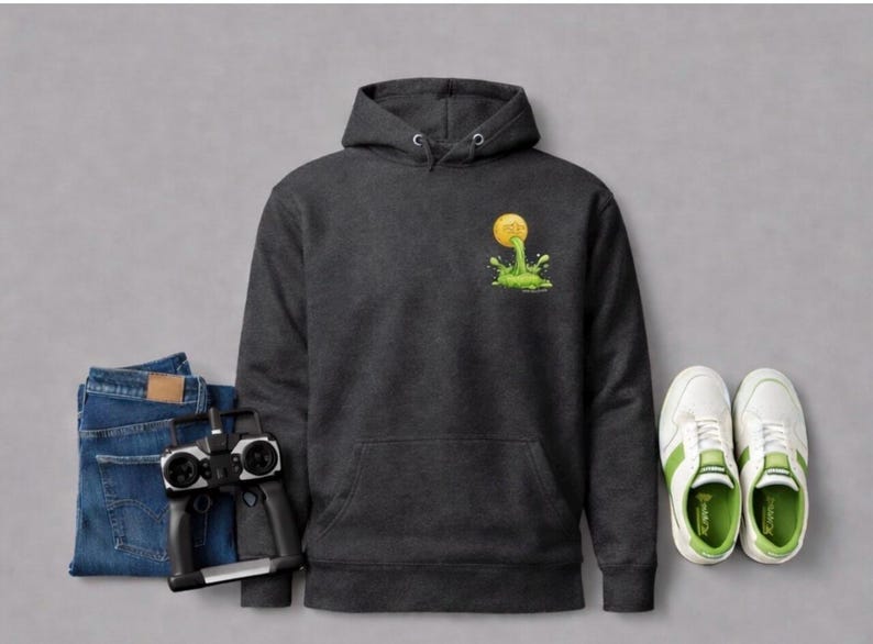 Peut inclure: Un sweat &agrave; capuche gris fonc&eacute; avec une poche avant et un graphisme d'une balle jaune avec de la slime verte. L'image comprend &eacute;galement un jean bleu, une t&eacute;l&eacute;commande et des baskets blanches avec des accents verts.