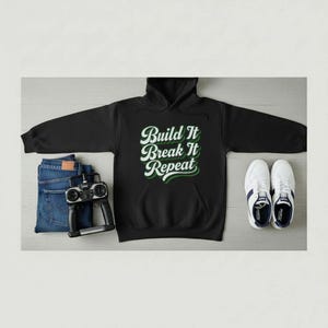 以下が含まれることがあります： 白と緑で「Build It Break It Repeat」の文字が入った黒いパーカー。画像には、青いジーンズ、リモコン、ネイビーブルーのアクセントが付いた白いスニーカーも含まれています。