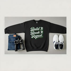 May include: Black sweatshirt with the text "Build It Break It Repeat" in white and green. The image also includes a pair of blue jeans, a remote control, and white sneakers with blue accents.