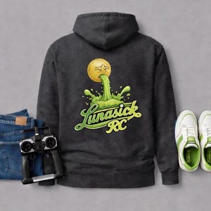 Peut inclure: Sweat &agrave; capuche gris fonc&eacute; avec un motif d'une balle jaune vomissant du liquide vert, avec le texte "Lunasick RC". L'image comprend &eacute;galement un jean bleu, une t&eacute;l&eacute;commande et des baskets blanches avec des accents verts.