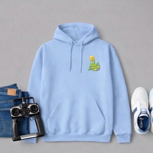 May include: A light blue hoodie with a front pocket and a small graphic on the chest. The graphic features a yellow flower and green text. The hoodie is paired with blue jeans, a remote control, and white sneakers.