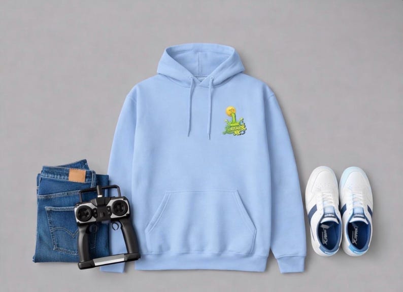 May include: A light blue hoodie with a front pocket and a small graphic on the chest. The graphic features a yellow flower and green text. The hoodie is paired with blue jeans, a remote control, and white sneakers.