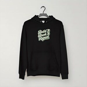 以下が含まれることがあります： フロントポケットとドローストリングフードが付いた黒いパーカー。パーカーには、白と緑のレトロフォントで「Build It Break It Repeat」というテキストが表示されています。パーカーは黒いハンガーに吊るされています。