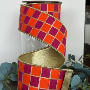 May include: Decorative ribbon with a vibrant orange and magenta checkered pattern, accented with gold trim. The ribbon is rolled and displayed, showcasing its design. The ribbon is suitable for crafts, gift wrapping, and decorative purposes.