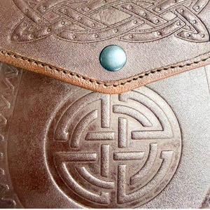 May include: Close-up of a brown leather pouch with Celtic knotwork and a circular design embossed on the front. The pouch has a flap secured by a blue button. The leather is textured and the stitching is visible along the flap.