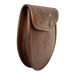 May include: A brown leather sporran with Celtic knotwork embossed on the flap and body. The sporran has a curved shape and a snap closure. The leather is a rich, medium brown color, and the stitching is visible along the edges.