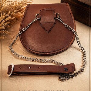 May include: A brown leather satchel with a silver chain strap and a separate leather belt. The satchel has a rounded shape and a flap closure. The text "HERITAGE IN EVERY DETAIL" is visible at the bottom of the image.