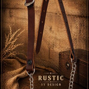 May include: A brown leather strap with a silver buckle and chain. The strap has a button closure and is connected to a silver chain. The text "RUSTIC BY DESIGN" is visible.