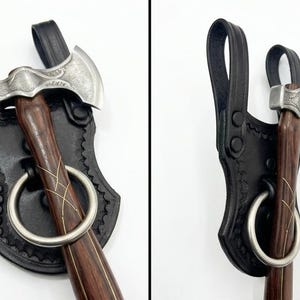 May include: A small, silver axe with a dark wooden handle and a decorative design. The axe is secured in a black leather sheath with a large metal ring. The sheath has a decorative edge and a loop for carrying.