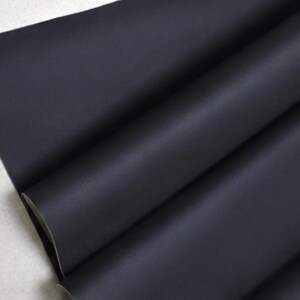 May include: Close-up of a folded piece of dark navy blue fabric. The fabric has a smooth, matte finish and is layered to reveal a white backing. The folds create shadows and add depth to the image, highlighting the texture and color.