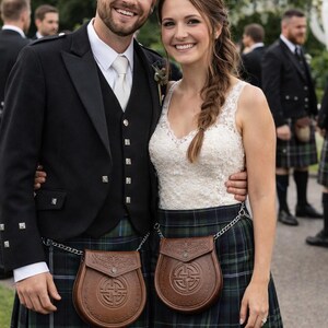 May include: A couple in traditional Scottish attire. The man wears a black jacket, vest, and kilt with a brown leather sporran. The woman wears a white lace top and a matching kilt. Both have brown leather sporrans with chain belts.