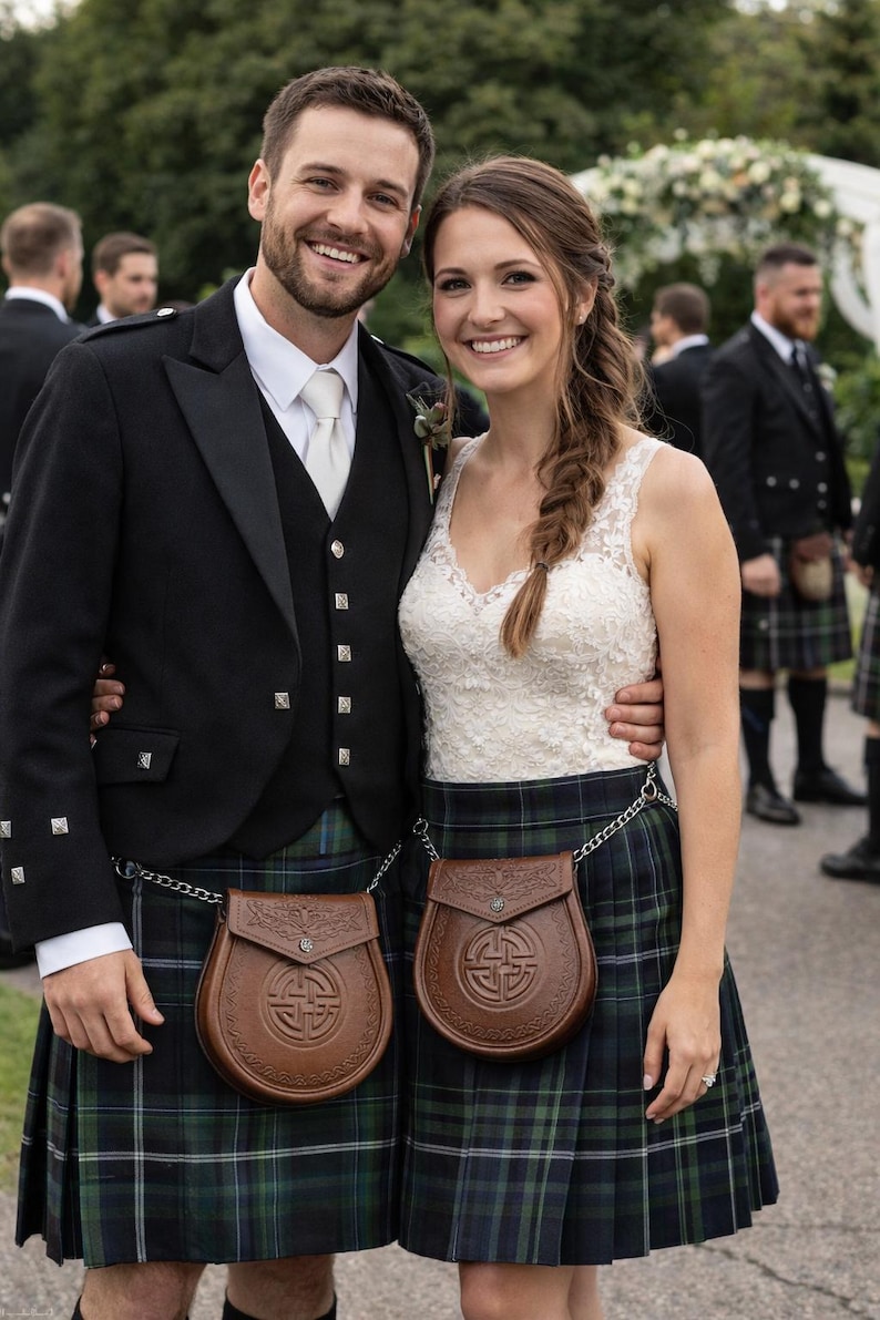 May include: A couple in traditional Scottish attire. The man wears a black jacket, vest, and kilt with a brown leather sporran. The woman wears a white lace top and a matching kilt. Both have brown leather sporrans with chain belts.
