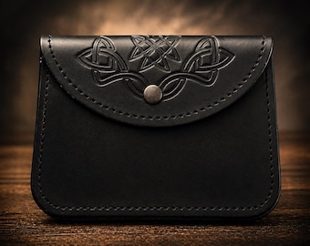 Peut inclure: Une pochette en cuir noir avec un motif de nœud celtique en relief sur le rabat. La pochette a une forme arrondie, une fermeture à bouton métallique et des coutures visibles. Le fond est une surface en bois flou.
