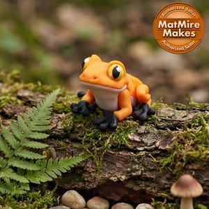 May include: A handcrafted orange frog figurine with large, expressive eyes and black feet, perched on a moss-covered log. The background features a fern, small stones, and a mushroom, creating a naturalistic scene. The text "MatMire Makes" is visible.
