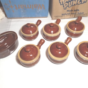 May include: A collection of brown ceramic soup bowls with lids and handles. The bowls have a light tan rim and a dark brown exterior. One larger oval-shaped casserole dish is also included in the set. The bowls are arranged on a white surface.