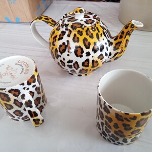 May include: A ceramic tea set featuring a leopard print design. The set includes a teapot and two matching mugs. The teapot has a lid and a curved spout. The mugs have handles. The color scheme is yellow, brown, and white.