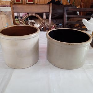 May include: Two antique stoneware crocks. One crock has a brown interior, the other has a black interior. Both are cylindrical with a light beige exterior and a rounded rim. They are sitting on a white surface.