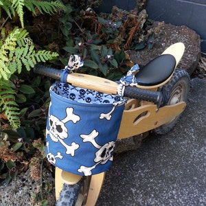 May include: A wooden balance bike with a blue and white skull and crossbones fabric basket attached to the handlebars. The bike has a black leather seat and black tyres.