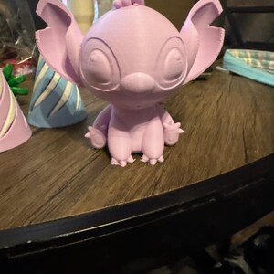 Puede incluir: Una figura de color p&uacute;rpura claro de un personaje de dibujos animados con orejas grandes, ojos grandes y brazos y pies peque&ntilde;os. La figura est&aacute; sobre una superficie de madera. Otros objetos son visibles en el fondo.