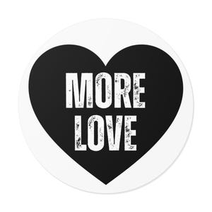 Könnte beinhalten: Ein runder Aufkleber mit einer soliden schwarzen Herzform auf weißem Hintergrund. Zentriert im Herzen befindet sich der Text MORE LOVE in einer fetten, weißen serifenlosen Schrift mit einer abgenutzten, verwitterten Textur.