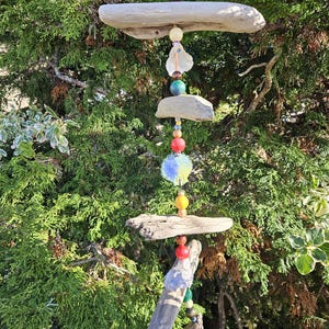 May include: A handcrafted wind chime featuring natural driftwood pieces and colorful beads. The chime includes various shapes and sizes of driftwood, interspersed with beads in shades of red, yellow, blue, and green. The wind chime hangs from a metal hook.
