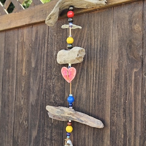 Puede incluir: Un móvil de viento artesanal de madera flotante con cuentas de colores y una pieza de cerámica en forma de corazón rosa. El móvil de viento también presenta una concha de ostra blanca en la parte inferior. Los elementos naturales crean una estética rústica y costera.