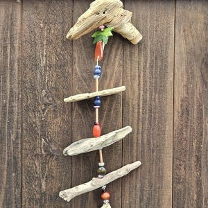 May include: A handcrafted driftwood mobile with natural wood pieces, colorful beads, and a small green leaf accent. The mobile hangs against a rustic wooden background, showcasing its organic design and earthy tones. The mobile is approximately 12 inches long.