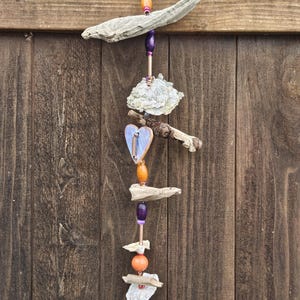 Puede incluir: Un móvil de viento artesanal de madera flotante con un acento en forma de corazón. El móvil presenta piezas de madera natural, cuentas moradas y naranjas, y un corazón azul claro. Cuelga de una valla de madera.