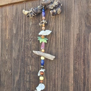 Puede incluir: Móvil artesanal de madera flotante y cuentas. Presenta piezas de madera natural, varias cuentas en tonos azules, amarillos y marrones, y elementos decorativos. Cuelga frente a una valla de madera.