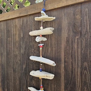 Puede incluir: Un móvil de viento de madera flotante con piezas de madera natural y cuentas de colores. El móvil cuelga de una cuerda azul y presenta varios tamaños de madera desgastada, conectados por cuerdas y cuentas. El fondo es una valla de madera.