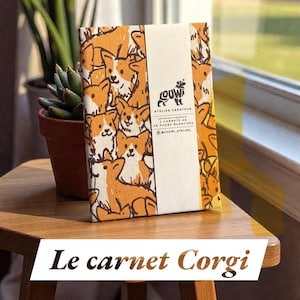 Pode incluir: Um caderno com um padrão repetido de ilustrações de corgis laranja e brancos. O caderno é embrulhado com uma faixa branca que inclui o texto "ATELIER CREATEUR" e "48 PAGES BLANCHES". O título "Le carnet Corgi" é exibido na parte inferior.