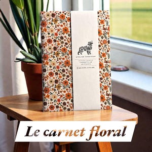 May include: A notebook with a floral pattern in shades of orange, brown, and cream. A white band with a logo and text wraps around the notebook. The text "Le carnet floral" is displayed at the bottom.