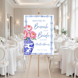 May include: A rectangular welcome sign on a wooden easel featuring a blue bamboo-style border, a blue and white porcelain vase with pink peonies, and the text: WELCOME TO Brunch WITH THE Bride IN HONOR OF Martha Harris MAY 23, 2026.