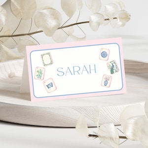 May include: A pink and white place card with the name "SARAH" in blue script. The card is decorated with watercolor illustrations of Mahjong tiles. The card is displayed on a white wooden tray.
