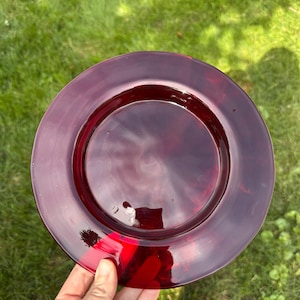 May include: A circular, translucent deep red glass plate with a smooth, polished surface and a wide, flat rim. The plate features a subtle, swirling texture within the glass material, creating a rich, dark crimson hue that catches the light.
