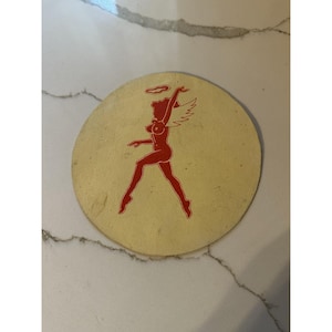 Puede incluir: Un posavasos redondo y beige con una silueta roja de una mujer con alas de ángel y un halo. La mujer está en una pose de baile. El posavasos tiene un aspecto ligeramente desgastado.