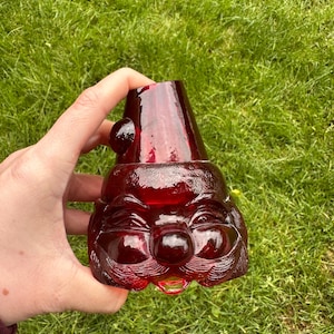 May include: A ruby red glass gnome-shaped container with a smiling face, round nose, and a conical hat. The container has a textured surface. It is held against a backdrop of green grass.