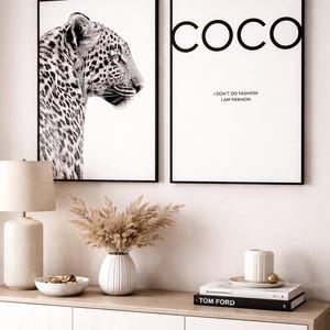 Puede incluir: Dos impresiones artísticas enmarcadas. Una presenta un retrato de leopardo en blanco y negro. La otra tiene la palabra "COCO" en letras negras grandes, con el texto "I DON'T DO FASHION I AM FASHION" debajo. Una lámpara, un jarrón con flores secas y libros están sobre un mueble blanco.