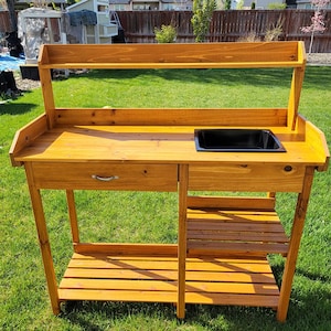 May include: A wooden potting bench with a natural finish, featuring a rectangular work surface, a built-in black plastic sink, a single drawer with a silver handle, an upper shelf, and a slatted lower storage shelf for gardening tools and supplies.