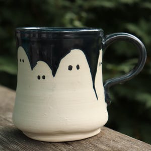 Peut inclure: Une tasse en céramique avec une base de couleur crème et un vernis bleu marine foncé qui coule du bord. La tasse présente un motif peint à la main de petites figures de fantômes simples avec deux points noirs pour les yeux, espacés.