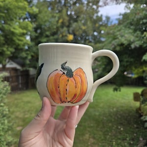 Peut inclure: Une tasse en céramique de couleur crème ornée d'une citrouille orange peinte à la main avec une tige vert foncé. La tasse présente un corps arrondi, une anse incurvée sur le côté droit et une finition lisse. Le motif inclut des contours noirs.