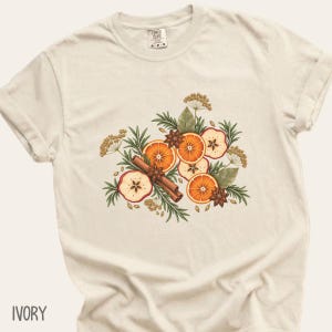 May include: An ivory short-sleeve t-shirt featuring a central graphic illustration of sliced oranges, dried apple slices, cinnamon sticks, star anise, and green sprigs. The shirt has a crew neckline and a Comfort Colors brand label at the neck.