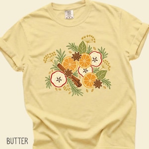May include: A butter yellow short-sleeve t-shirt featuring a central graphic illustration of sliced oranges, apple rounds, cinnamon sticks, star anise, and green sprigs. The shirt has a crew neckline and a label at the inner collar.