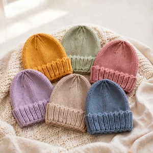 Può includere: Una collezione di sei berretti in maglia in vari colori a tinta unita, tra cui giallo senape, verde salvia, rosa antico, lavanda, beige e blu ardesia. Ogni berretto presenta un risvolto a coste e una lavorazione a maglia rasata.