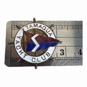May include: A circular metal pin shaped like a ship's wheel with a white enamel border reading TAMAQUA YACHT CLUB. The centre features a red and blue burgee flag with a white zigzag stripe. A metal ruler shows the pin is 3.8 cm in diameter.