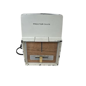 Puede incluir: Una tostadora Proctor-Silex vintage con exterior blanco y panel de imitación madera. La tostadora tiene un panel de control con ajustes para tostadas y pasteles. El nombre de la marca está impreso en la parte superior.