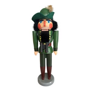 May include: A wooden nutcracker figurine in a green uniform and hat, with a black beard and binoculars. The figurine has a painted face with rosy cheeks and a stern expression. The base is grey.