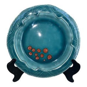 May include: A teal ceramic bowl with a wavy rim and a smooth, deep center. The bowl features a pattern of curved lines around the edge and small, orange-red circles in the center. It is displayed on a black stand.