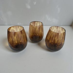 May include: Three stemless, oval-shaped drinking glasses. The glasses are a warm amber colour with a mottled pattern. The base of each glass is a darker shade of brown, creating a gradient effect. The glasses are likely used for beverages.